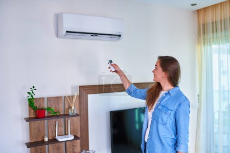 Foto de Joven hembra ajustando la temperatura del aire acondicionado en el control remoto en casa en la sala de estar - Imagen libre de derechos