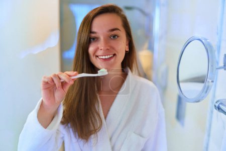 Foto de Young beautiful happy girl wearing soft white bathrobe brushing her teeth in bathroom. Care oral health - Imagen libre de derechos