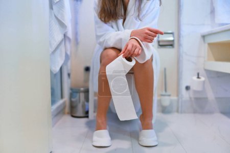 Photo for Female holding toilet paper roll and sitting on toilet in restroom - Royalty Free Image
