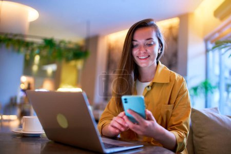 Photo for Happy cute joyful modern satisfied smart young casual girl freelancer wearing wireless headphones using phone and laptop for remotely working in a cafe - Royalty Free Image