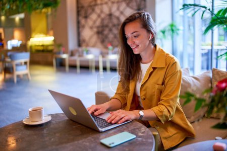 Happy cute joyful modern satisfied smart young casual girl freelancer wearing wireless headphones works remotely at laptop in a cafe