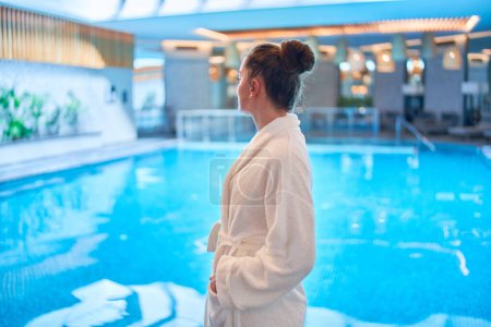 Carefree serene female wearing bathrobe chilling and relaxing alone at swimming pool in spa center. Mind health and getaway