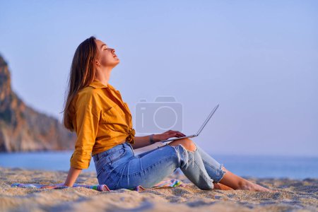 Zufriedene Millennial Freelancer Frau mit Computer und am Sandstrand am Meer sitzen. Genuss des Traum-Büro-Fernarbeits-Konzepts