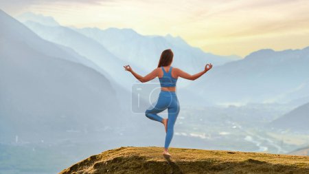 Téléchargez les photos : Vue arrière du calme calme gratuit serein heureux satisfait femme de remise en forme faisant de l'exercice de yoga sur le rocher supérieur contre idyllique vue sur la montagne du matin. Soins mentaux et habitudes saines - en image libre de droit