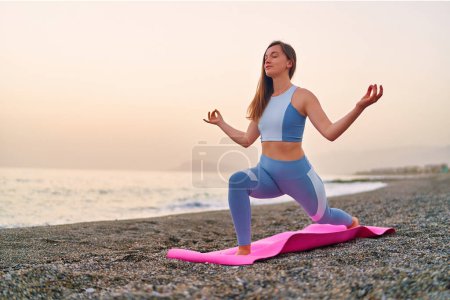 Téléchargez les photos : Calme bonheur serein femme de remise en forme satisfaite faisant méditation de yoga sur tapis et exercices de respiration sur la plage au bord de la mer. Soins mentaux et habitudes saines - en image libre de droit