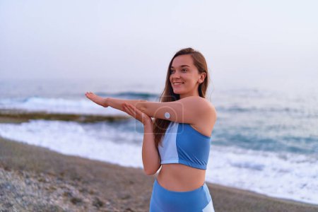 Téléchargez les photos : Satisfait une femme diabétique mince de forme physique faisant des exercices d'étirement des mains par la mer. Habitudes saines et mode de vie actif diabétique - en image libre de droit