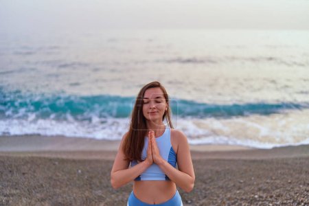 Foto de Mujer fitness con los ojos cerrados y las manos orantes de pie solo con la posición de meditación mientras se respiran ejercicios por mar. Cuidado mental y hábitos saludables - Imagen libre de derechos