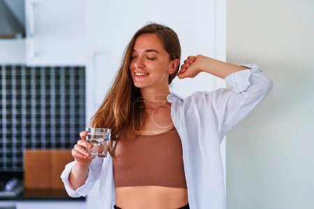 Téléchargez les photos : Portrait de jeune heureuse satisfaite jolie femme jolie boire un verre d'eau pure filtrée fraîche le matin après le réveil. Commencez la journée avec des habitudes saines et soutenez l'équilibre quotidien de l'eau - en image libre de droit