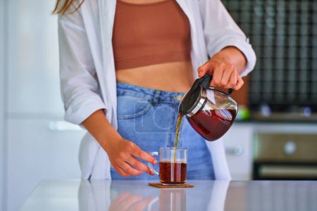 Photo for Female pouring black tea from a glass teapot into a cup at home kitchen - Royalty Free Image