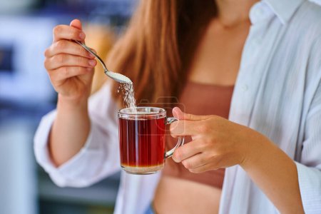 Téléchargez les photos : Mains féminines versant cuillère à sucre dans une tasse en verre avec du thé noir chaud - en image libre de droit