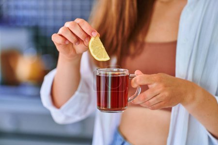 Téléchargez les photos : Femme buvant du thé noir chaud avec une tranche de citron à la cuisine maison - en image libre de droit