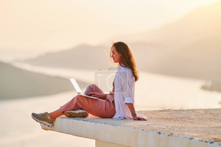 Foto de Inspirado hembra feliz sentado en la parte superior y disfrutando de distancia remota en línea de trabajo en el ordenador con hermosa vista al atardecer - Imagen libre de derechos
