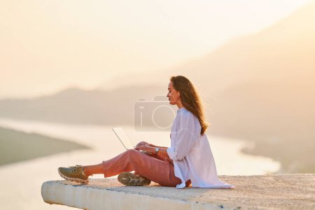 Téléchargez les photos : Inspiré heureux femelle assis sur le dessus et profiter de la distance de travail en ligne à distance à l'ordinateur avec une belle vue au coucher du soleil - en image libre de droit