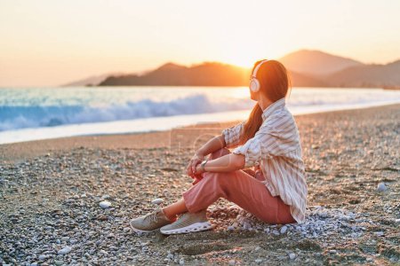 Foto de Mujer tranquila sentada sola y escuchando relajarse viajando música en la orilla del mar al atardecer. Hermoso momento de vida vacaciones - Imagen libre de derechos