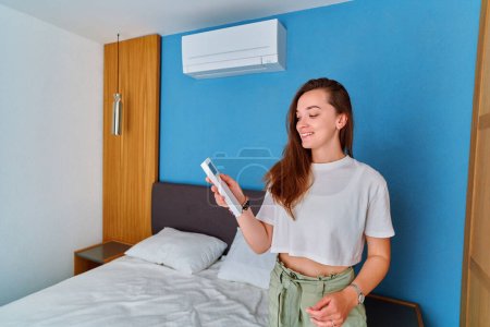 Foto de Joven linda chica sonriente ajusta la temperatura del aire acondicionado utilizando control remoto en el dormitorio - Imagen libre de derechos