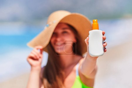 Téléchargez les photos : Femme appliquant et tenant bouteille vide blanche vide de crème solaire tout en bronzant sur la plage au bord de la mer par une journée d'été ensoleillée. Protection solaire - en image libre de droit