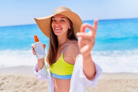 Téléchargez les photos : Joyeuse souriante joyeuse femme caucasienne insouciante tenant une bouteille vide blanche vide de crème solaire sur la plage de la mer par une journée d'été ensoleillée. Protection solaire - en image libre de droit