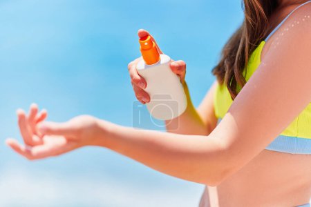 Téléchargez les photos : Femme appliquant et tenant bouteille vide blanche vide de crème solaire tout en bronzant sur la plage au bord de la mer par une journée d'été ensoleillée. Protection solaire - en image libre de droit