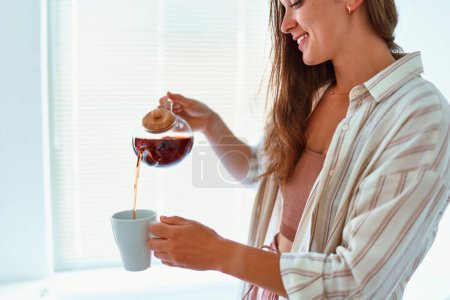 Téléchargez les photos : Heureux jeune femme joyeuse et souriante heureuse versant du thé chaud dans une tasse de théière en verre à la maison - en image libre de droit