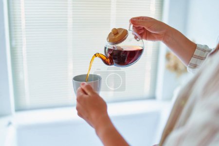 Foto de Mujer vertiendo té negro aromático caliente en la taza de la tetera de vidrio en casa - Imagen libre de derechos