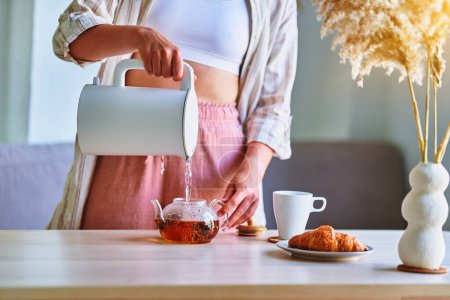 Téléchargez les photos : Femme versant de l'eau bouillante dans une tasse de bouilloire électrique moderne pour préparer le thé à la maison - en image libre de droit