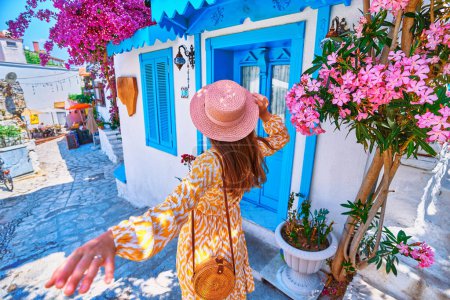 Foto de Sígueme concepto. Chica viajero vistiendo vestido y sombrero camina por la hermosa calle de flores de colores con casas blancas y puertas azules en una ciudad europea - Imagen libre de derechos