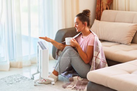 Photo for Happy smiling satisfied joyful young woman with plaid and knitted socks drinking hot tea and warming near portable electric heater at home - Royalty Free Image