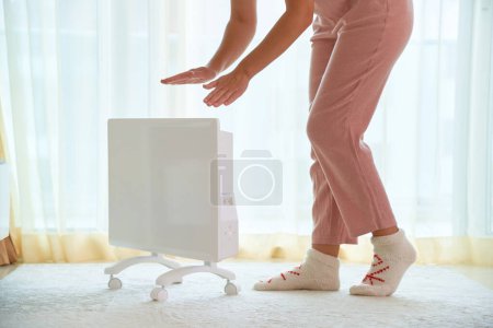 Foto de Mujer con calcetines de punto suave comodidad acogedora obtener calentamiento manos sobre moderno calentador eléctrico portátil blanco en casa - Imagen libre de derechos