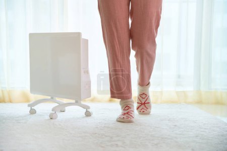 Foto de Mujer con calcetines de punto suave comodidad acogedora obtener calentamiento con moderno calentador eléctrico portátil blanco en casa - Imagen libre de derechos
