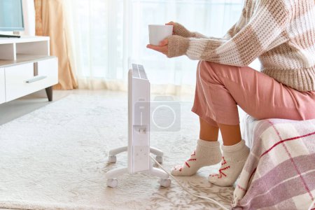 Foto de Acogedora mujer que usa calcetines de punto y ropa suave comodidad obtener calentamiento con calentador eléctrico portátil blanco moderno en casa - Imagen libre de derechos