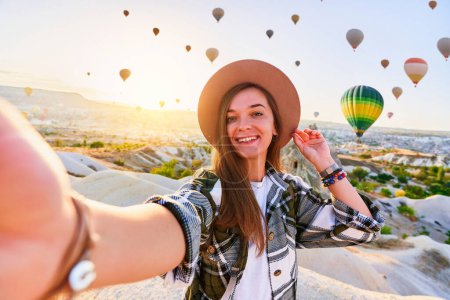 Photo for Happy smiling joyful satisfied girl traveling blogger takes selfie photo in Nevsehir, Goreme. Beautiful destination with colorful flying hot air balloons in Anatolia, Kapadokya - Royalty Free Image