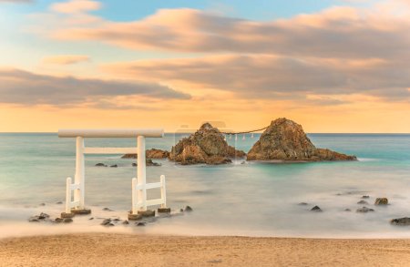 Fotografía del paisaje marino de larga exposición de la playa de Itoshima y sus famosas rocas de pareja Meoto Iwa de Sakurai Futamigaura protegidas por una puerta torii Shinto blanca sagrada en la luz del atardecer.