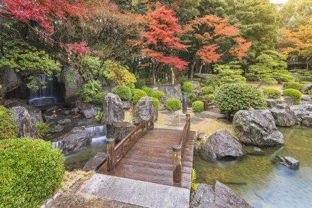 Foto de Fukuoka, kyushu - december 07 2022: The Nuno-ochi falls crossed by a wooden bridge surrounded by rocks and overlooked by red momiji maple trees in the Japanese Ohori garden a rainy afternoon. - Imagen libre de derechos