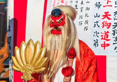 Téléchargez les photos : Tokyo, setagaya - january 28 2023: Japanese man dressed in Suzukake kimono wearing a Tokin headwear and a bearded red mask of Tengu holding a golden fan during the Shimokitazawa parade festival. - en image libre de droit