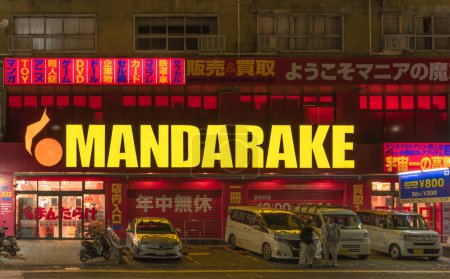 Foto de Fukuoka, japan - dec 07 2022: Facade of the Mandarake store specialize in manga and anime-related and adorned by a giant neon sign lighting in the night above the cars parked on its parking area. - Imagen libre de derechos