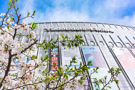 Foto de Tokyo, japan - march 29 2022: Low-angle shot of Japanese sakura cherry blossoms blooming in front of the facade of the Department store Yodobashi-Akiba specialized in home appliance in Akihabara. - Imagen libre de derechos