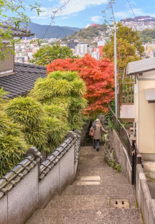 Téléchargez les photos : Une femme âgée descend une pente raide avec un escalier étroit descendant les collines de Nagasaki entre les maisons japonaises dont la clôture permet aux arbustes de bambou et aux érables momiji rouges de faire saillie en automne - en image libre de droit