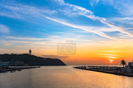 Photo for Kanagawa, enoshima - dec 25 2022: Sunset on the Enoshima island topped by the Enoshima Sea Candle tower with the fishing port of katase in foreground and the Sakai river flowing into the Sagami bay. - Royalty Free Image
