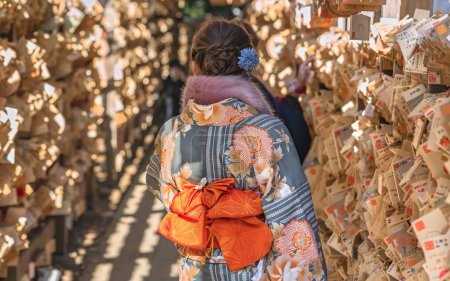 Téléchargez les photos : Femme japonaise de retour portant un kimono d'hiver avec des motifs de fleurs, une ceinture obi et une écharpe en fourrure avec une coiffure de chignon et une épingle à cheveux fleur de soie entourée de tablettes de prière en bois shinto ema. - en image libre de droit