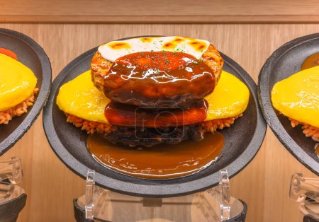Téléchargez les photos : Modèle alimentaire japonais ou échantillon de shokuhin représentant une omelette d'omurice faite avec du riz frit et surmontée d'un steak de hambourg avec du fromage gruyere fondu et une sauce brune, le plat préféré des enfants. - en image libre de droit
