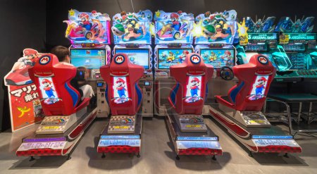 Foto de Tokyo, Japón - 22 ago 2023: Asientos deportivos electrónicos iluminados alineados con un jugador sentado jugando al juego Mario Kart Arcade GP DX desarrollado por Nintendo y Bandai Namco en un centro de juegos japonés. - Imagen libre de derechos