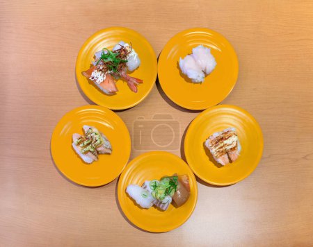 Foto de Fotografía de ángulo superior de un surtido de nigiri-zushi o sushi en cinco platos amarillos dispuestos en círculo sobre la mesa de un restaurante Kaiten-zushi con camarones, vieira, salmón, calamar, pulpo o atún - Imagen libre de derechos