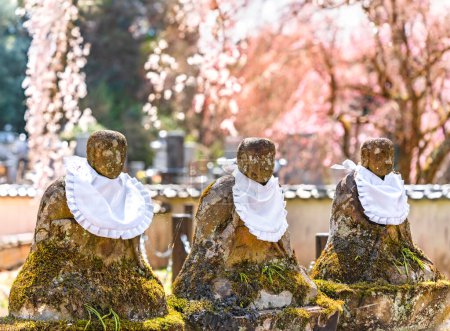 Foto de Tres estatuas de piedra de Jizo Bodhisattva o Jizo Bosatsu conocido como protector de niños nacidos muertos cubiertos de musgo centenario y con un babero sobre un fondo bokeh de flores de cerezo rosa - Imagen libre de derechos