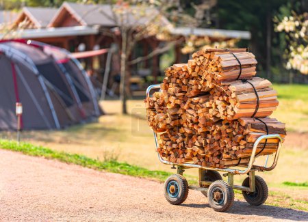 Focus on a small metal cart with large rubber wheels heavily loaded with a bunch of bales of pieces of firewood on a sunny path leading to a campsite with a tent on the lawn in the background.