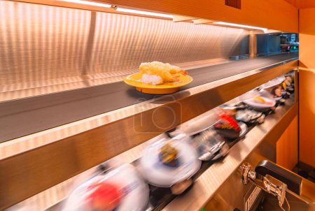 Langzeitbelichtungsfotos einer umtriebigen, sich drehenden Sushi-Bar, die die Essenz des schnellen japanischen Essens mit einem Teller frittiertem Shrimp Tempura Sushi einfängt, lieferten das oben genannte Förderband.