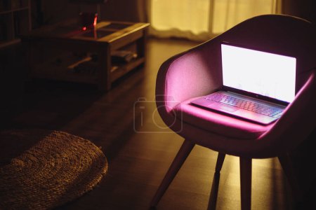 Photo for Dimly lit dining room where there is a table and a chair with a laptop lit with dim and colorful light that dyes the environment with light - Royalty Free Image