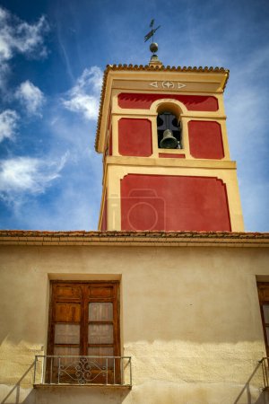 Der kastanienbraune Glockenturm des Parroquia de San Bartolome in Librilla, Murcia, Spanien ragt hinter der Fassade eines alten Hauses hervor