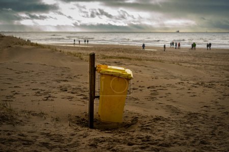 Téléchargez les photos : Yellow plastic trash can on the sand beach and people walking on the seashore, Katwijk, Netherlands - en image libre de droit