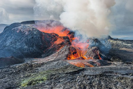 Foto de Vapor pesado y humo sobre un cráter volcánico. lava roja brillante fluye de un cráter activo. Paisaje en Islandia en la península de Reykjanes. Cielos nublados a la luz del día y el sol - Imagen libre de derechos