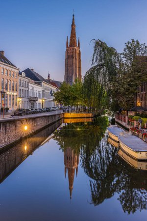 Photo for Canal in Bruges in the evening. Historic houses and the Church of Our Lady in the center of the old town of the Belgian Hanseatic city. Boats and trees with footpath and road - Royalty Free Image